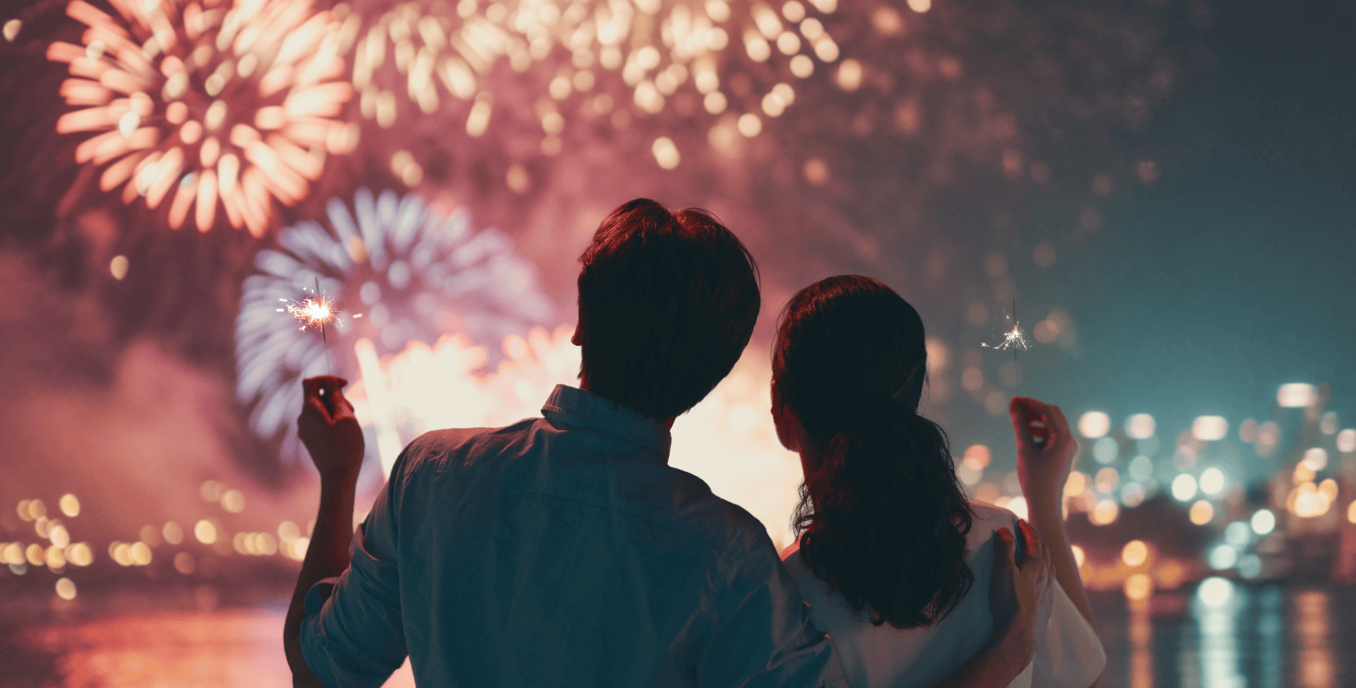 Two people stand close together at night, each holding a sparkler, watching colorful fireworks illuminate the sky over a cityscape across the water during Chinese New Year celebrations.