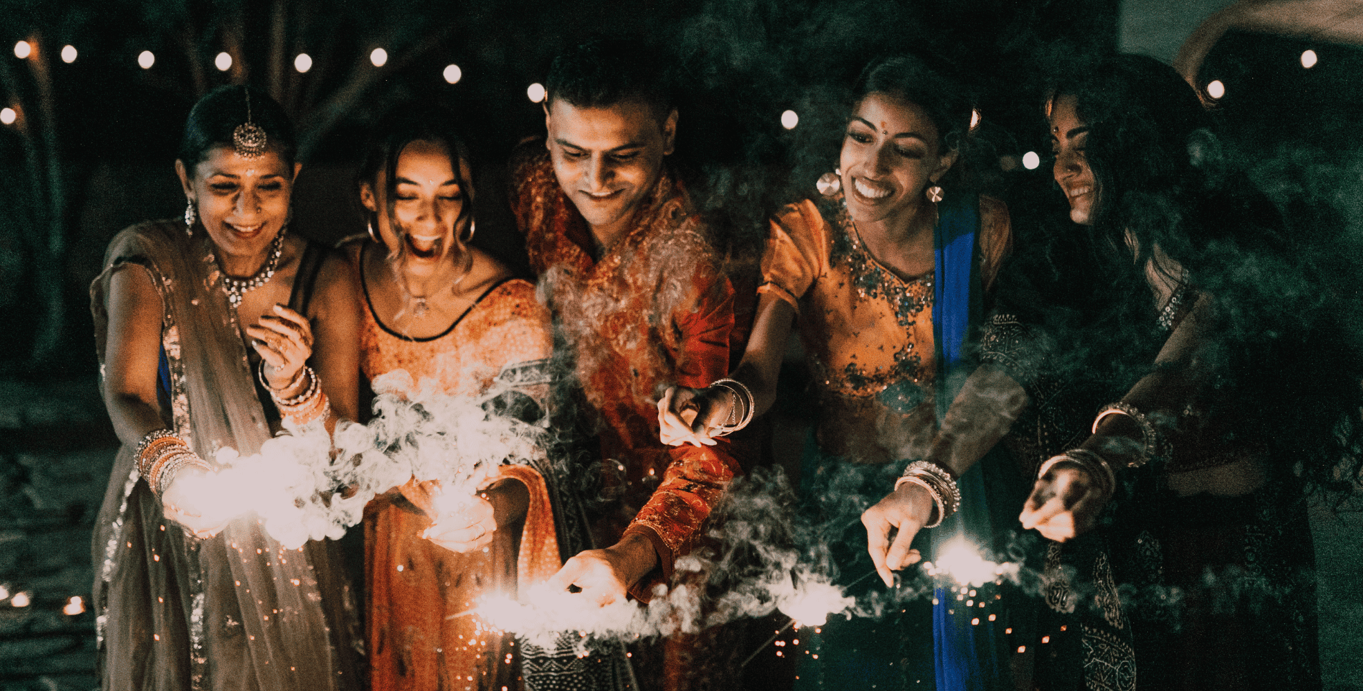 A group of people in festive attire hold sparklers, smiling and standing close together. Bright lights and smoke surround them, creating a celebratory atmosphere.