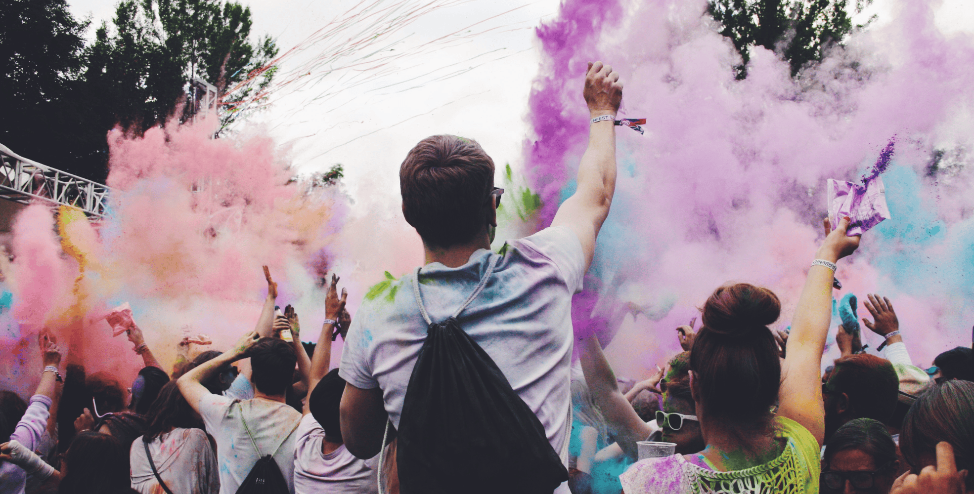 A crowd of people celebrate at an outdoor festival, throwing colorful powder into the air.