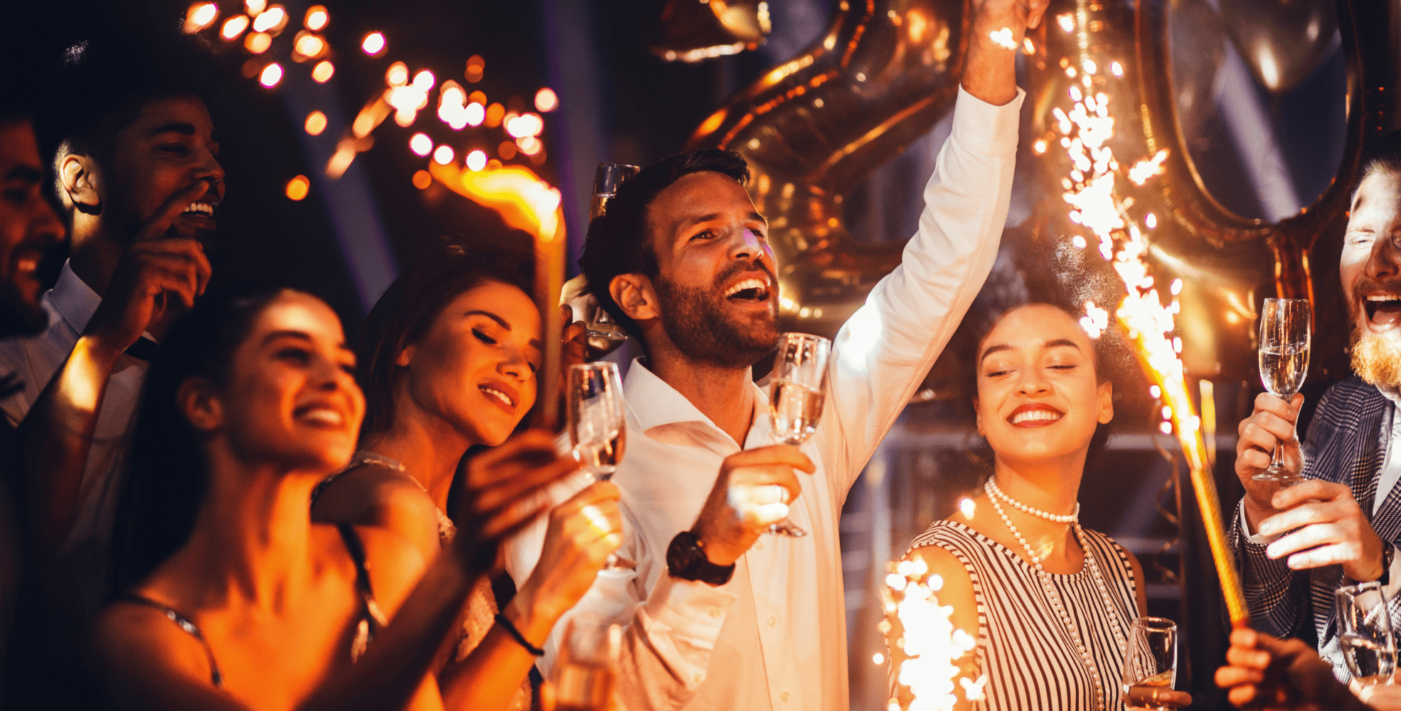 A group of people celebrating at a New Year's Eve party, holding champagne glasses, with sparklers and balloons in the background.