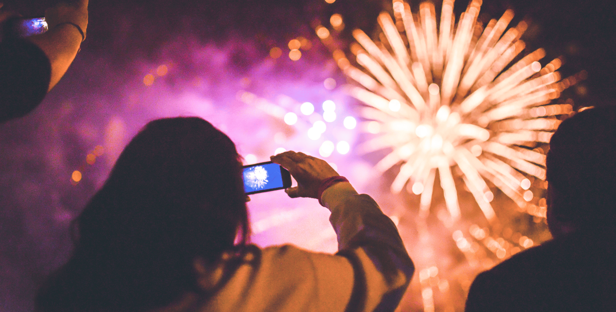 Amid lively celebrations, a person captures bright fireworks in the night sky with a smartphone, surrounded by others watching the dazzling display.