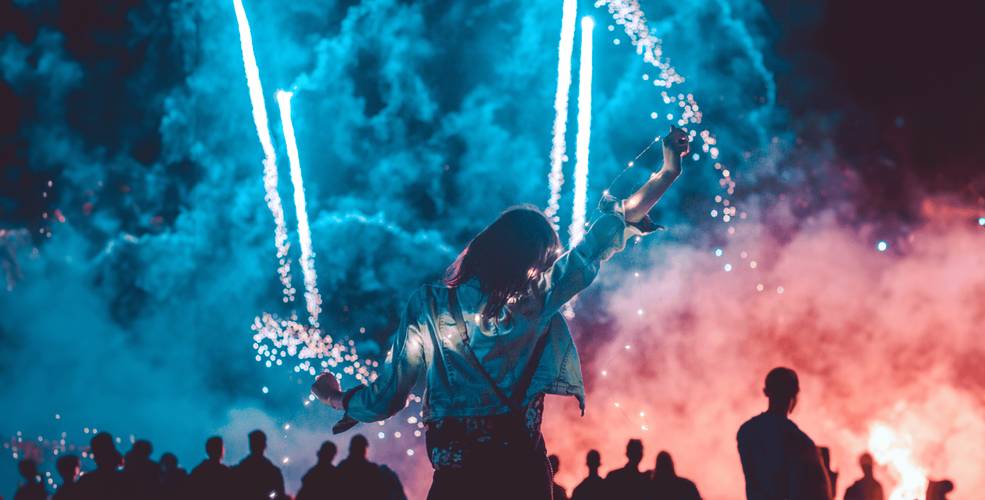 Person raises arms toward colorful fireworks and smoke in the night sky, with a crowd of silhouetted people in the foreground.