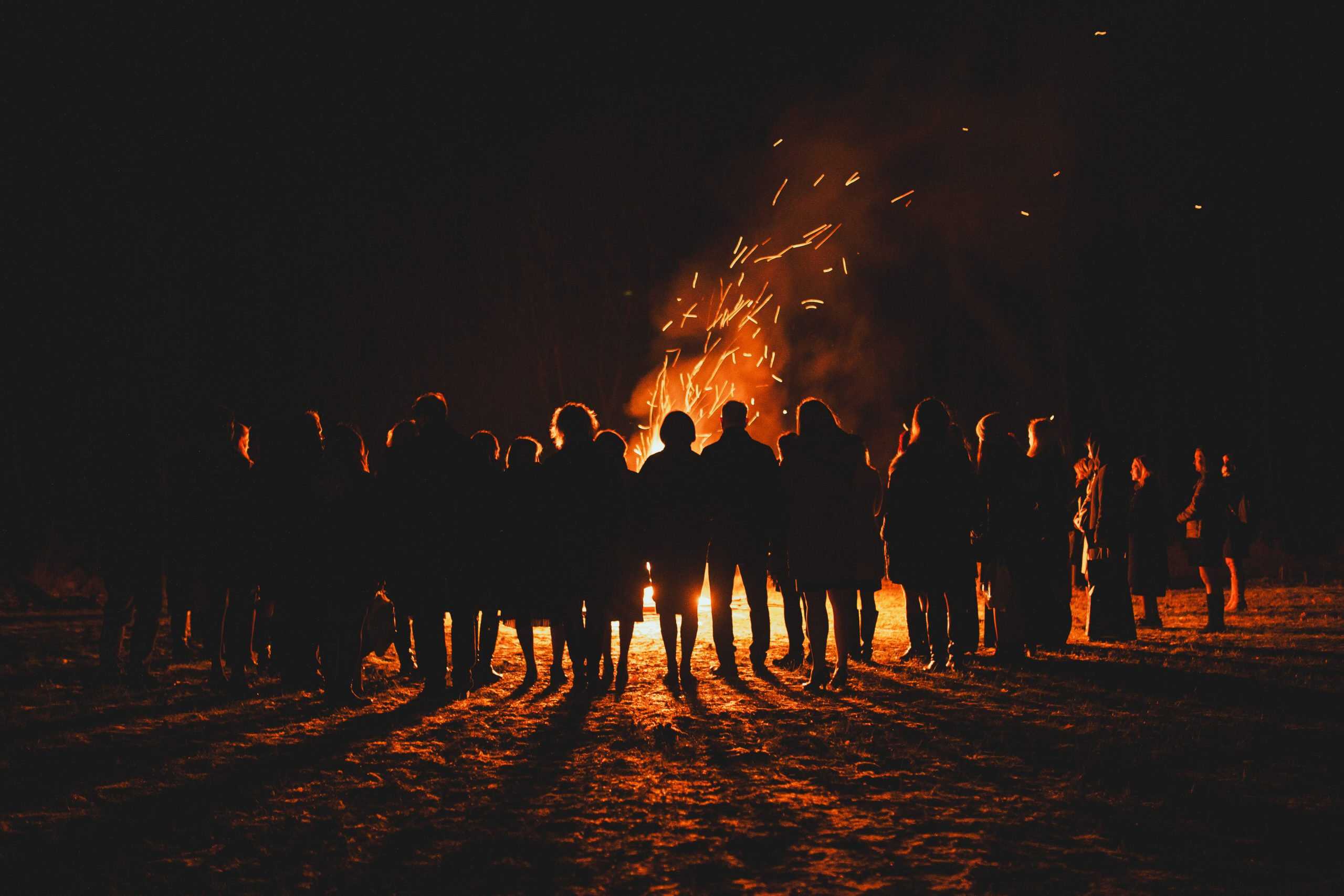 Silhouettes of people gathered around a large bonfire on Bonfire Night, with sparks and fireworks illuminating the dark sky.