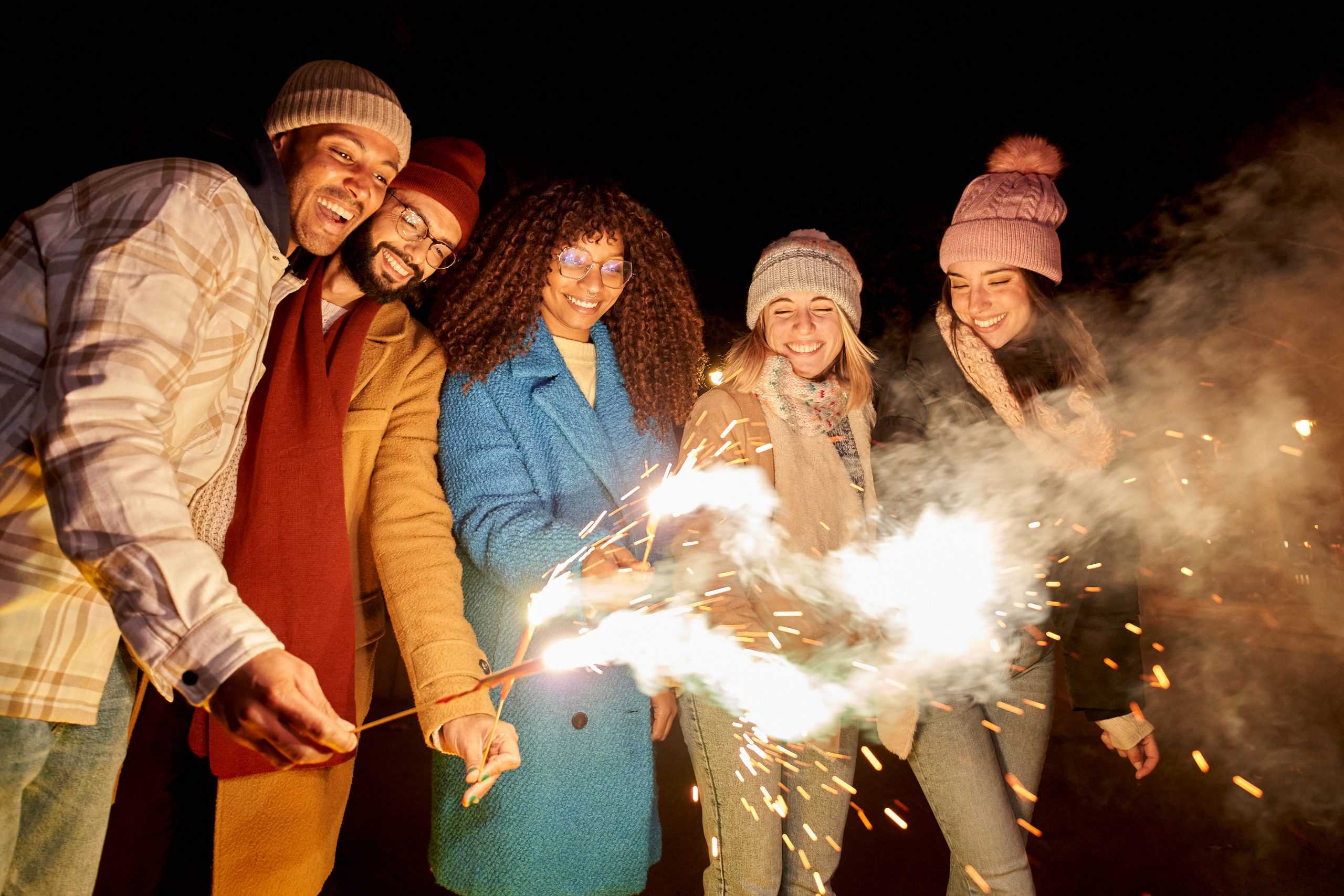 Five people in winter clothing hold sparklers, their faces lit with joy, as fireworks burst in the night sky during a Bonfire Night celebration.