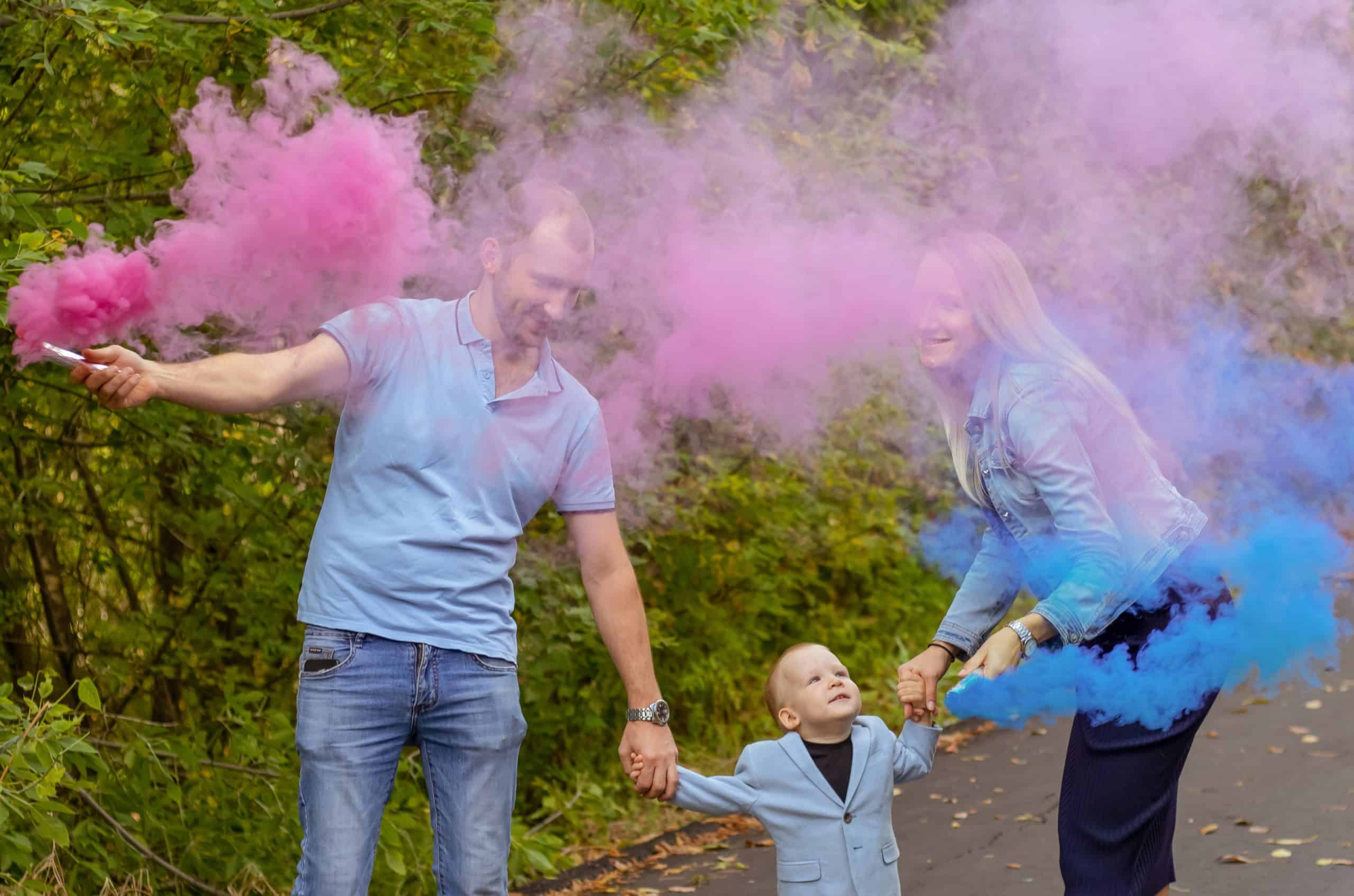 Two adults and a child stand on a path surrounded by trees, holding colored smoke sticks emitting pink and blue smoke—a joyful gender reveal celebration.