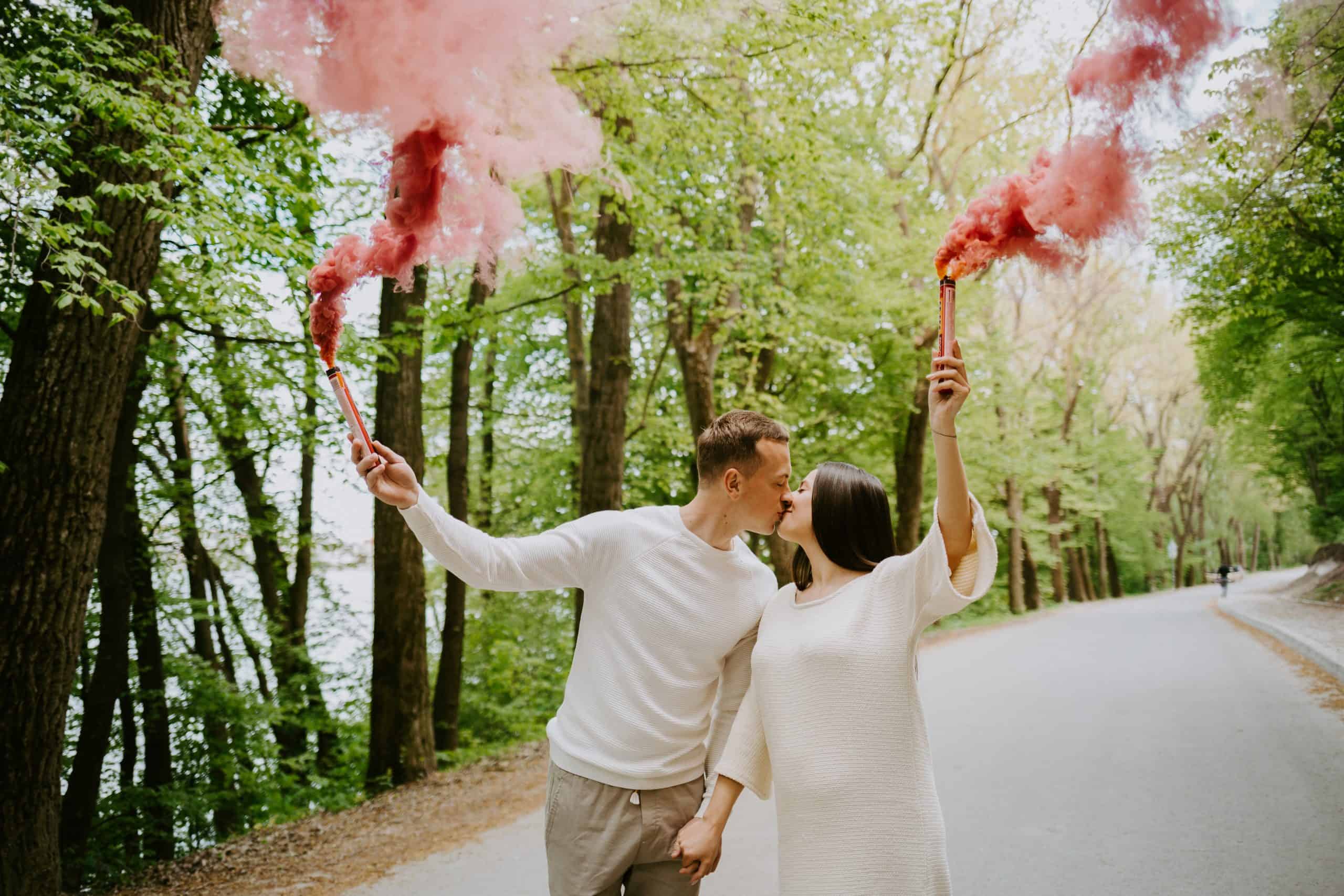 A couple shares a kiss, holding red smoke flares like a vibrant announcement in the forested roadside setting, reminiscent of a dramatic gender reveal.