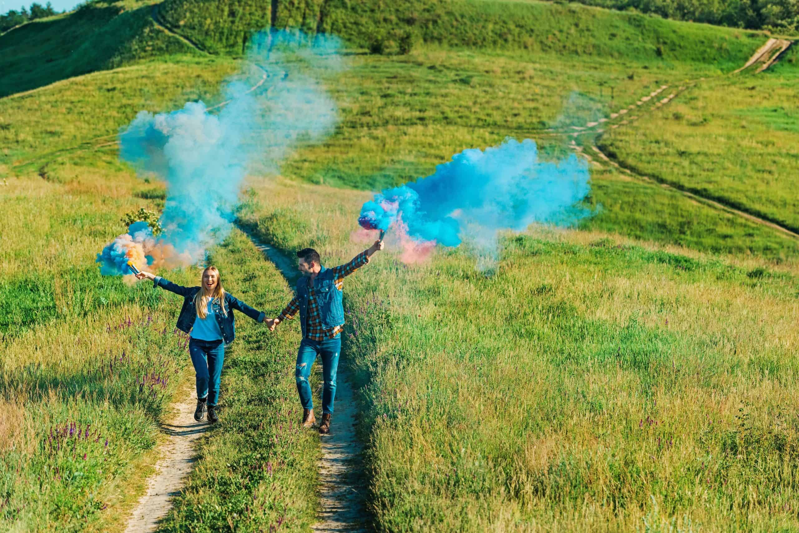 Two people hold hands, walking on a grassy path with colored smoke bombs for a gender reveal. The background features green hills under a clear sky.