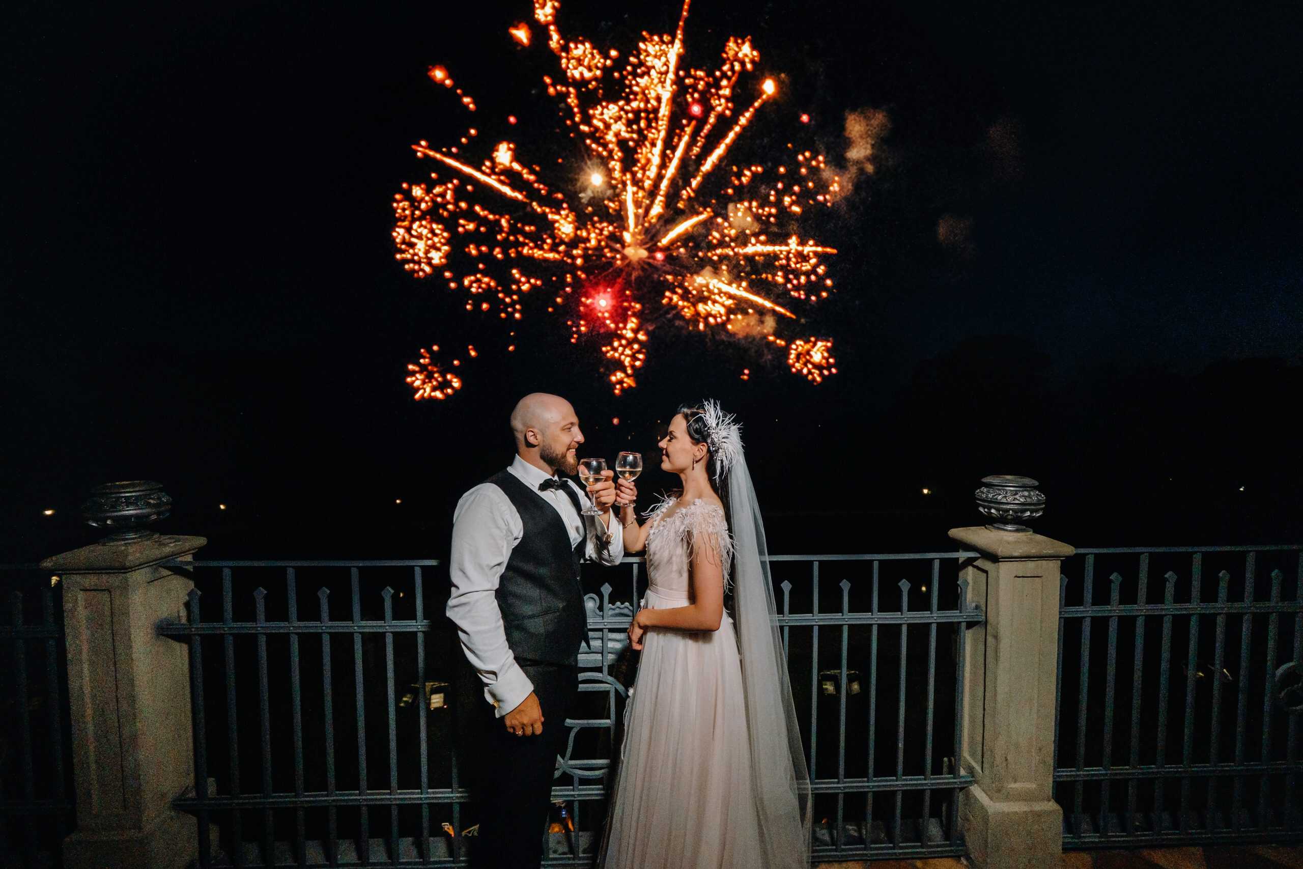 A bride and groom toast with champagne on a balcony at night, with fireworks lighting up the sky in the background.