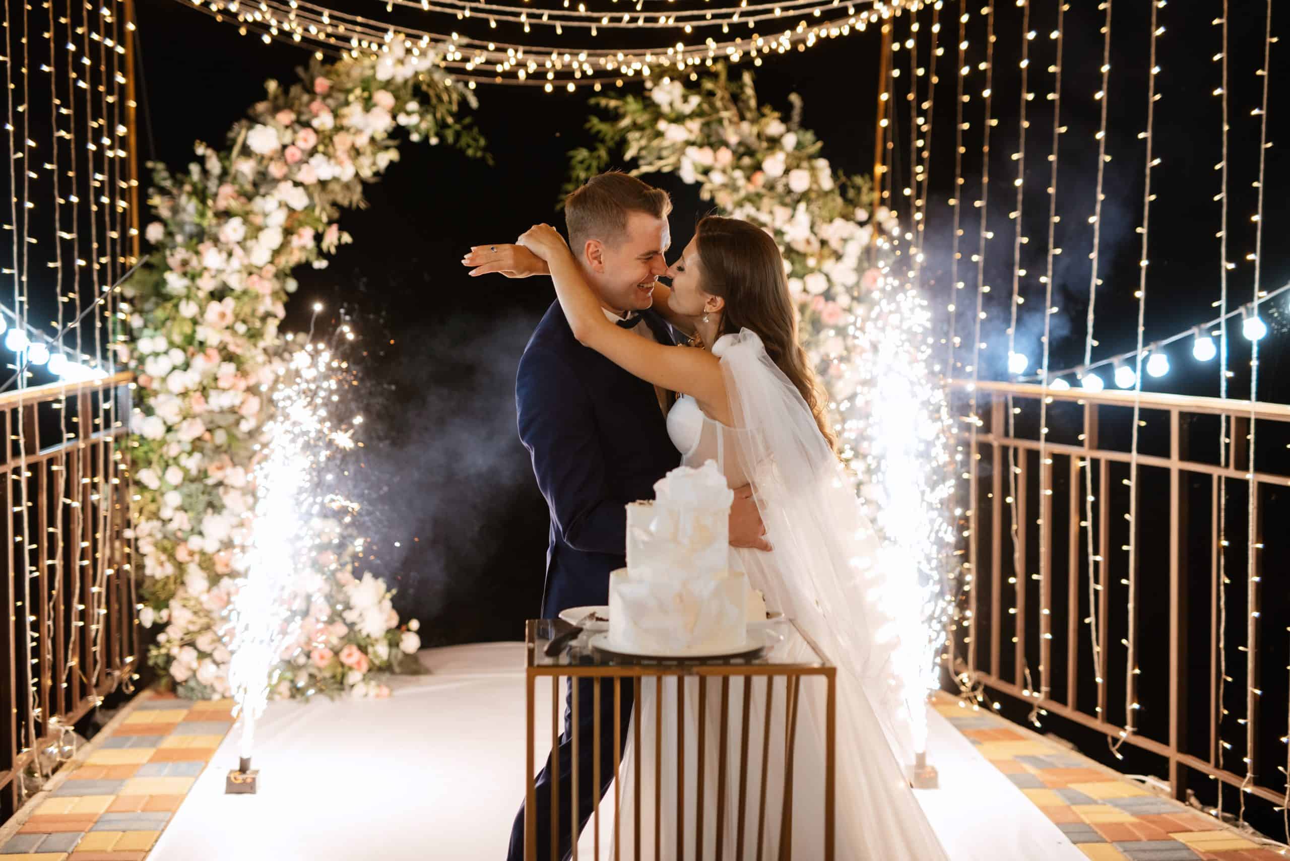 A couple embraces in front of a decorated archway with flowers and lights. There is a lit cake on a stand, and sparklers are going off in the background.