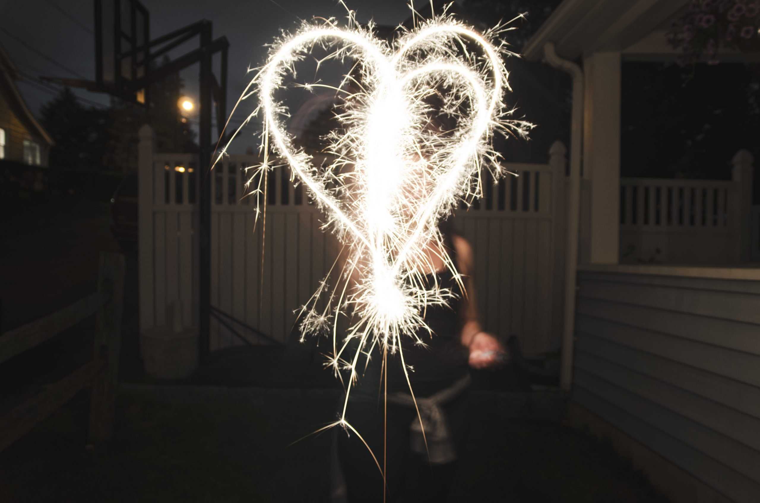 A heart shape drawn with a sparkler at night in front of a person. A fence and house are visible in the background.
